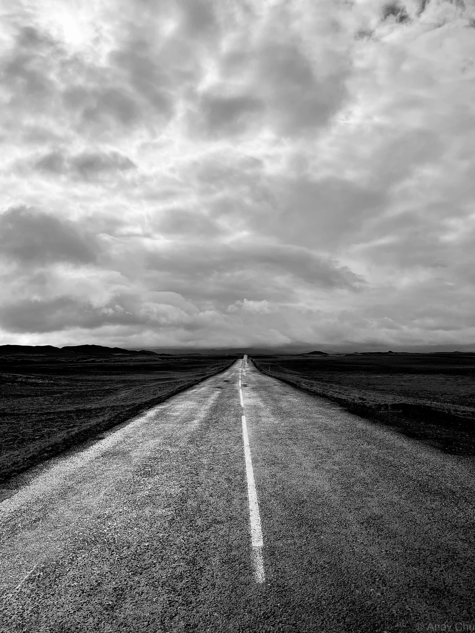 Black-and-white road stretching toward distant hills beneath clouds