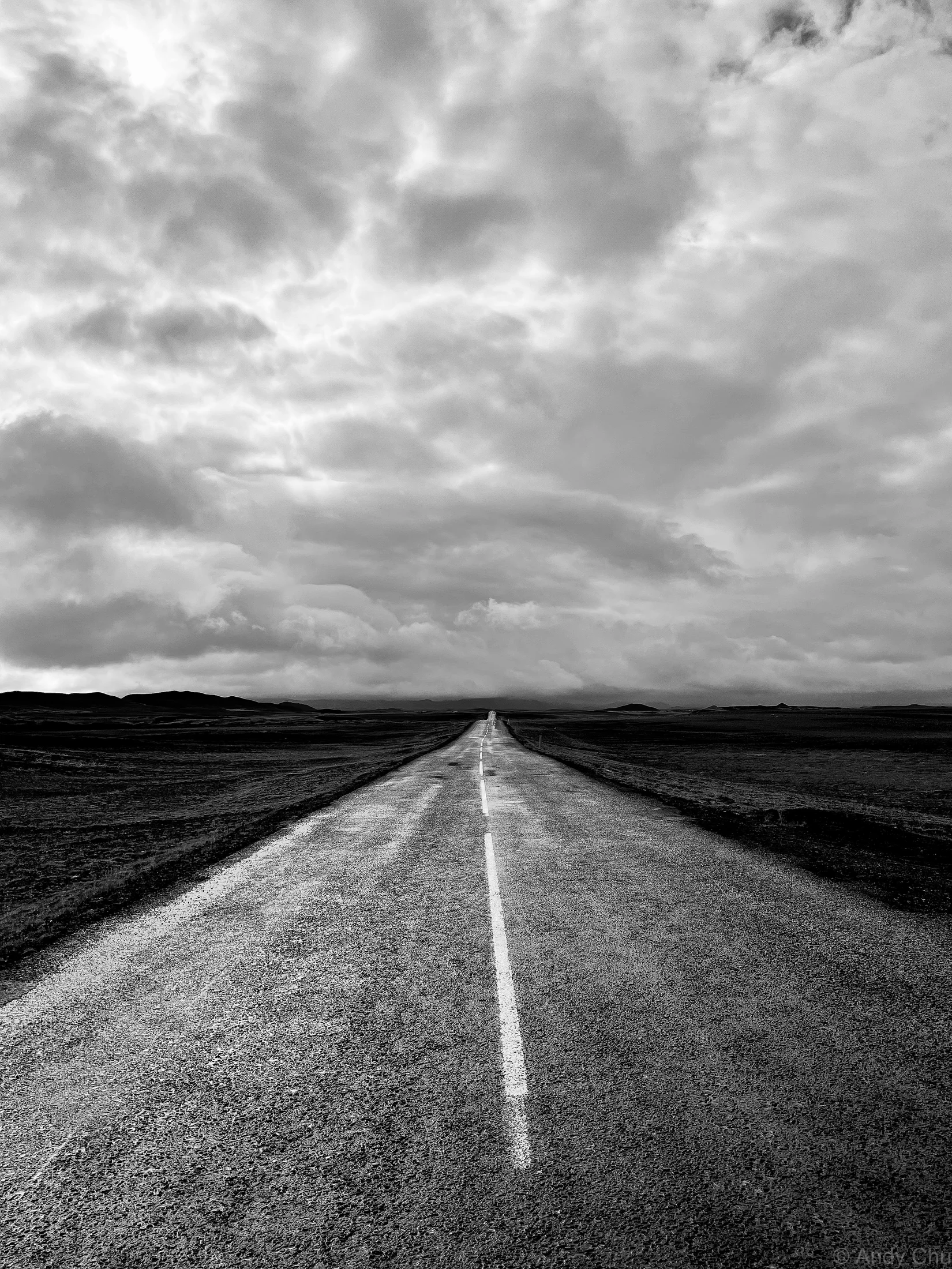 Black-and-white road stretching toward distant hills beneath clouds