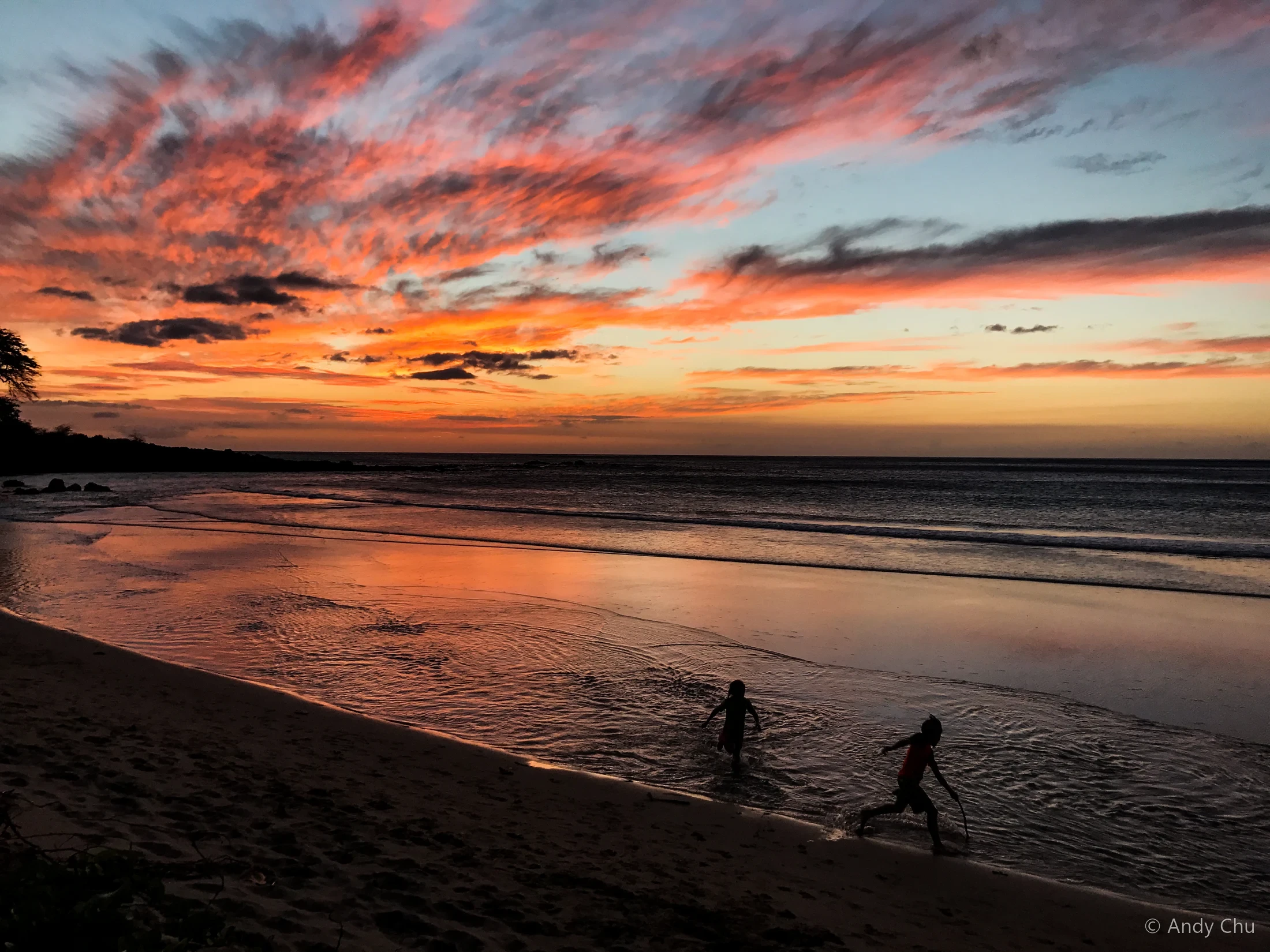 Two small figures playing at the water’s edge beneath a colourful sunset