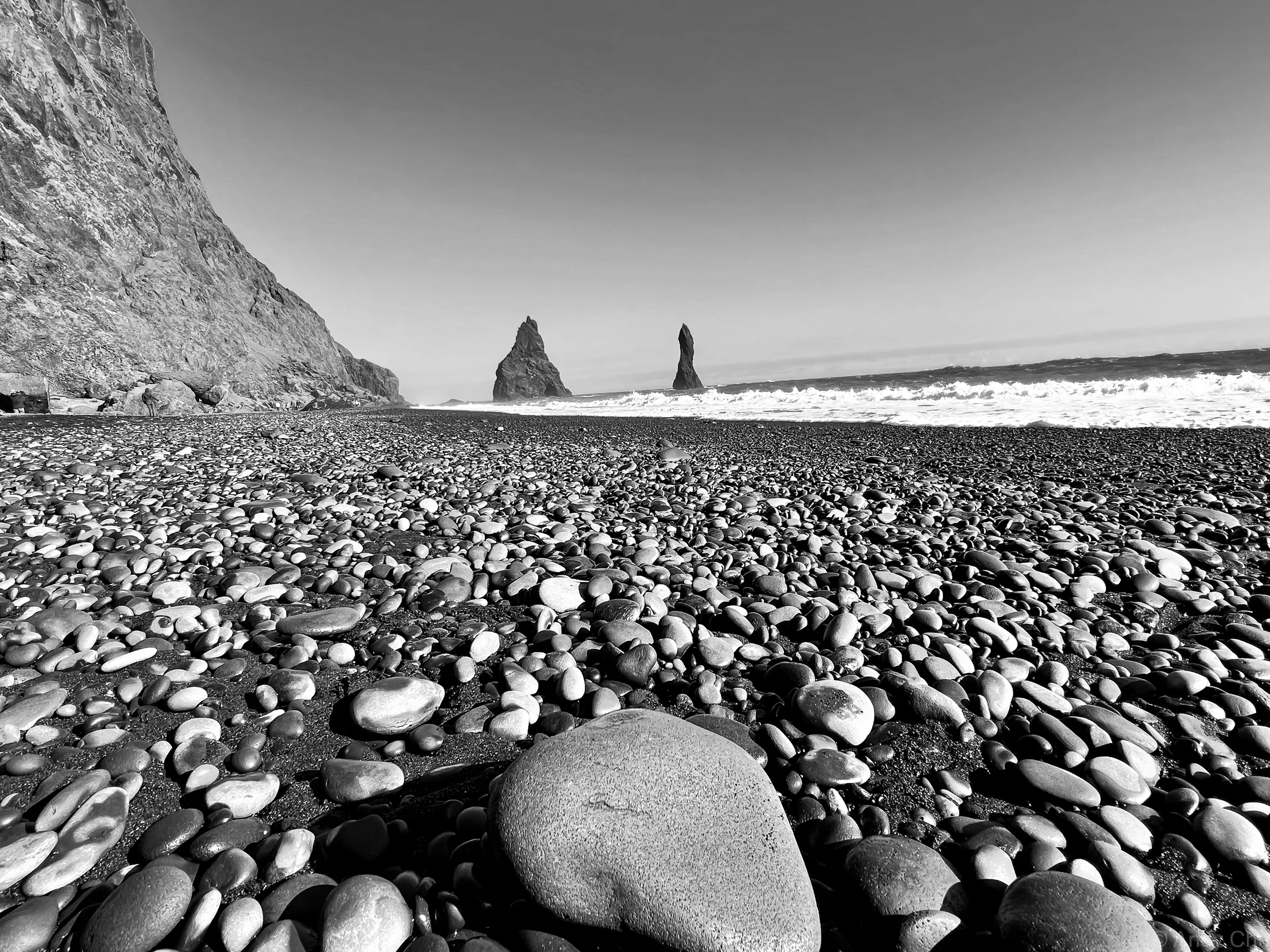 Black-and-white view across a pebble beach toward sea stacks
