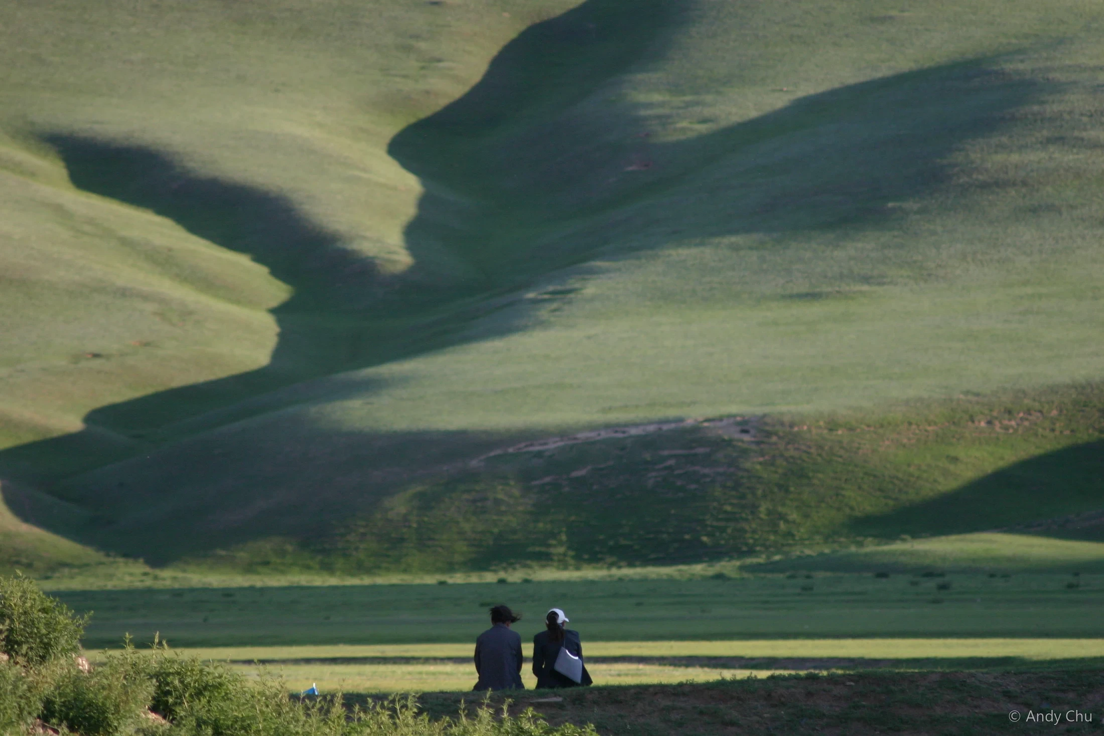Two figures in a wide green valley with shadowed hills