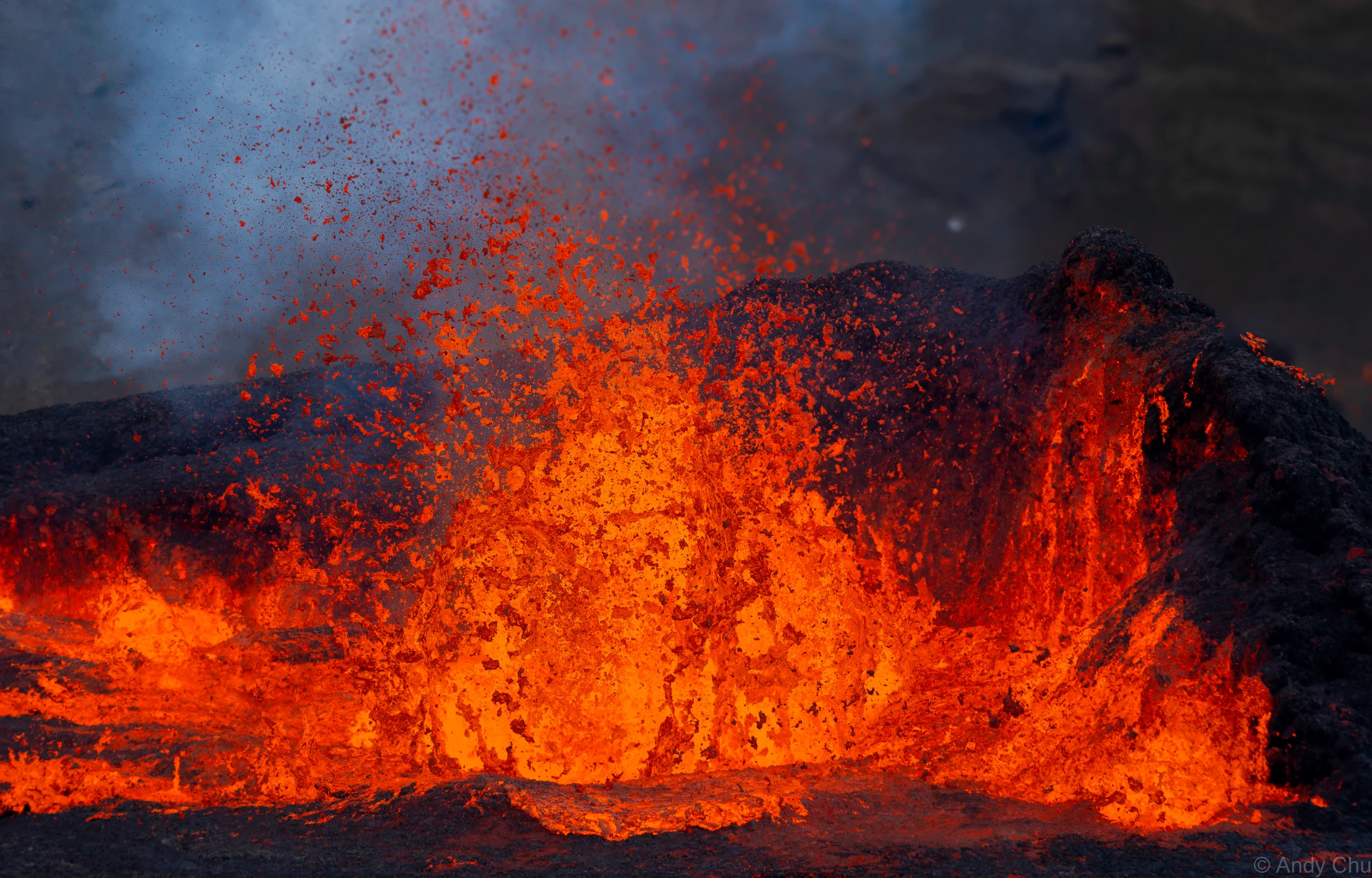 Orange lava erupting against dark rock and a grey sky